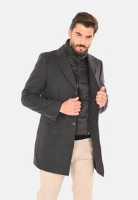 Uomo che sistema un soprabito grigio scuro indossato sopra una giacca trapuntata nera e pantaloni beige su uno sfondo chiaro e uniforme.