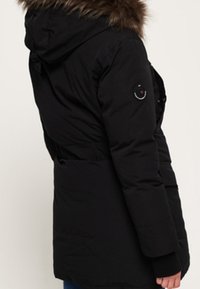 Svart isolerad parka-jacka med en huva klädd i päls, sidofickor och en figurnära design. Har en logotyp på ärmen.