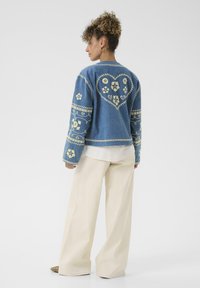 Denimska jakna s rumenim cvetličnim vezenjem, na hrbtu ima dizajn srca, dolge rokave in sproščen kroj, v kombinaciji s širokimi bež hlačami.