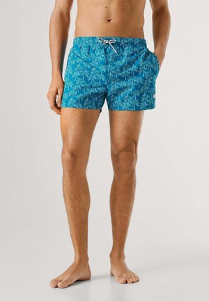 Man in blauwe zwemshorts met wit palmboompatroon, blootsvoets staand tegen een effen lichte achtergrond.