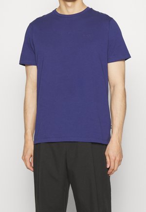 Homme portant un t-shirt uni bleu foncé à manches courtes et un col rond, ainsi qu'un pantalon noir, debout devant un fond clair.