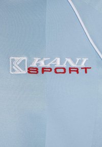 Šviesiai mėlyna sportinė medžiaga su tekstūriniu paviršiumi; turi išsiuvinėtą "KANI" baltos spalvos ir "SPORT" raudonos spalvos. Papuošta balta apdaila.