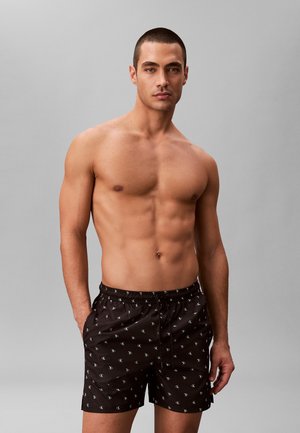 Homme musclé aux cheveux courts portant un short de bain noir Calvin Klein, debout devant un fond gris uni, les mains dans les poches.