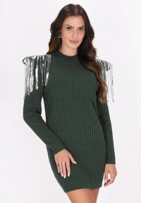 Abito maglione verde a costine con maniche lunghe, caratterizzato da dettagli in frange argentate sulle spalle. Realizzato in tessuto testurizzato, dalla vestibilità aderente.