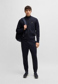 Tuta navy in tessuto liscio con giacca con zip, polsini a costine e pantaloni coordinati. Presenta un dettaglio con logo dorato sul petto.
