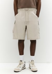 Pantaloni cargo beige con vita elasticizzata, cordino, tasche laterali e una vestibilità comoda, abbinati a sneakers alte bianche e beige.