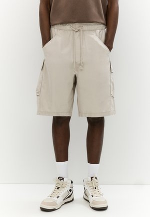 Shorts cargo beiges avec une taille élastique, cordon de serrage, poches latérales et une coupe décontractée, associés à des baskets montantes blanches et beiges.