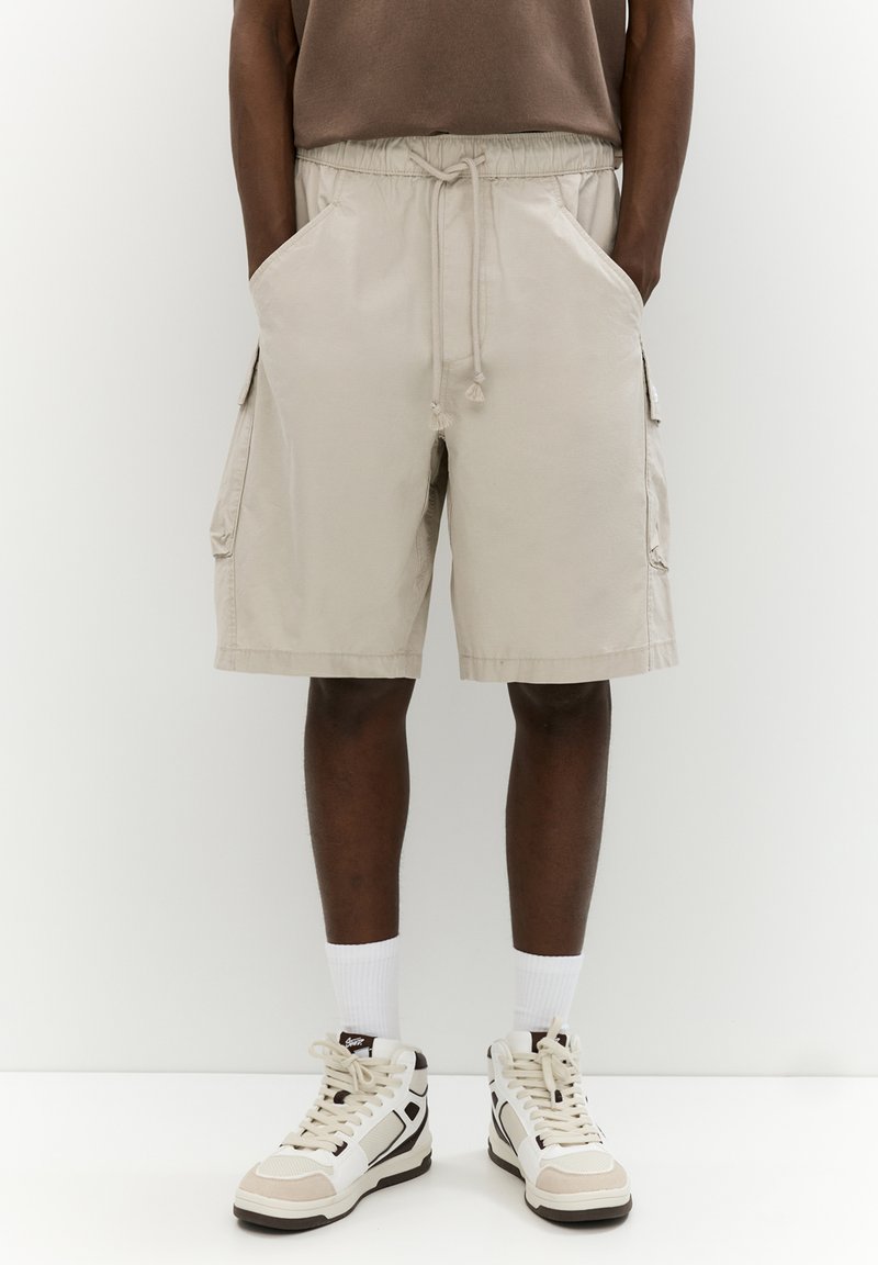 Pantaloni cargo beige con vita elasticizzata, cordino, tasche laterali e una vestibilità comoda, abbinati a sneakers alte bianche e beige.