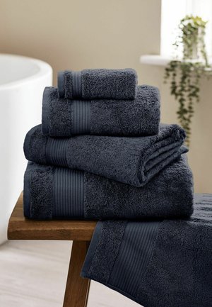 Mucchio di asciugamani piegati di colore grigio scuro di varie dimensioni posizionati su una panca in legno vicino a una vasca da bagno con una pianta sospesa sullo sfondo.