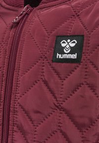 Quiltad vinröd jacka med dragkedja, prydd med en svart logotyppatch med en bi och varumärket "hummel". Strukturerat diamantmönster över hela.