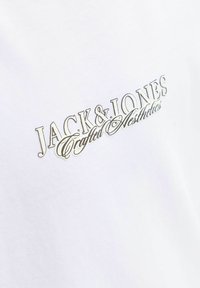 Witte katoenen t-shirt met een metallic zilveren logo tekst "JACK & JONES" met cursieve accenten boven "Crafted Aesthetics." Glad textuur.