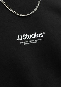 Fekete anyagú póló, fehér "JJ Studios" felirattal és szlogennel. Egy ezüst lánc nyaklánc fémes részletet ad. Az anyag sima textúrájú.