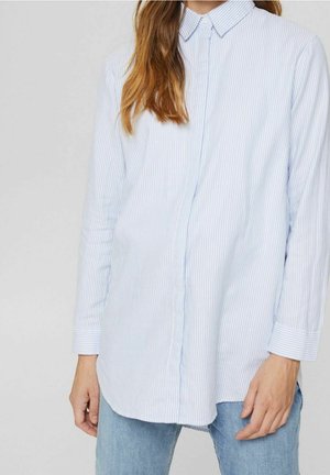 Kobieta ubrana w jasnoniebieską i białą cienką koszulę na guziki z długim rękawem w paski, w niebieskich dżinsach, stojąca na jednolitym tle.