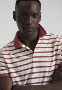 Polo a rayas en color crema con rayas rojas, con cuello rojo y mangas cortas, hecho de una tela texturizada y con cierre de botones en la parte delantera.