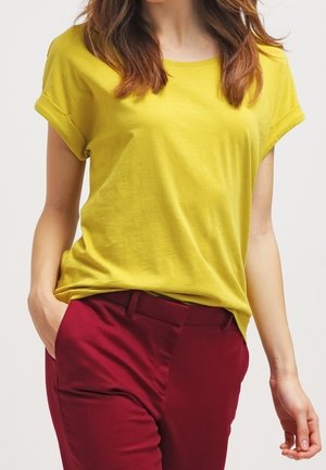 Kvinde iført gul kortærmet T-shirt stoppet ned i højtaljede mørkerøde bukser, den ene hånd i lommen, foran en ensfarvet baggrund.