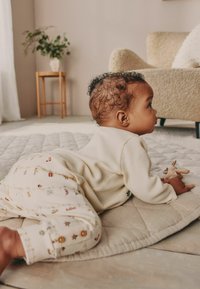 Kremo spalvos megztinis su ilgomis rankovėmis ir raštuoti pižamos džinsai. Kūdikiui poilsiaujant ant šviesiai iškloto kilimėlio, rankose laikomas mažas žaislinis žaislas.