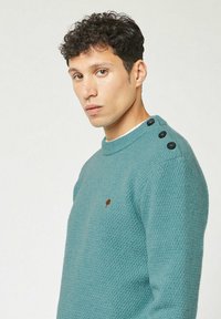 Junger Mann mit lockigem Haar, der einen strukturierten türkisfarbenen Pullover mit drei schwarzen Knöpfen an der Schulter und einem kleinen braunen Logo auf der Brust trägt.