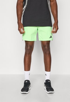 Groene sportshorts met een gladde textuur, voorzien van een zwart Adidas-logo en contrasterende zwarte strepen aan de zijkanten. Gekocht met zwarte sneakers.