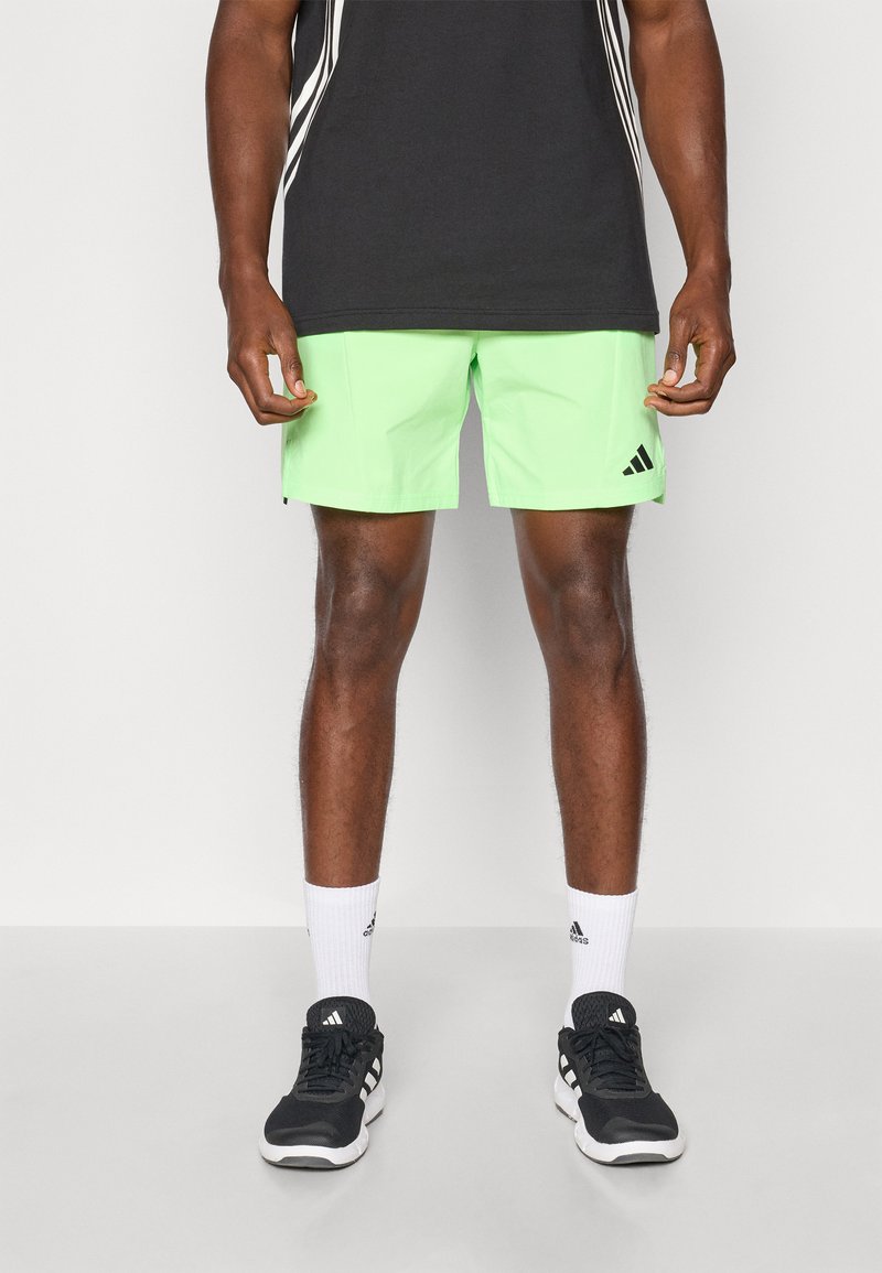 Shorts de sport verts avec une texture lisse, arborant le logo Adidas noir et des rayures noires contrastantes sur les côtés. Portés avec des baskets noires.