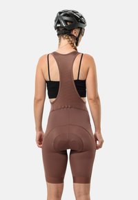 Bruine fiets-bibshorts met een racerback ontwerp, met een gevoerde zit en elastische pijpen. Draagt een zwarte helm, getoond vanuit de achterkant.