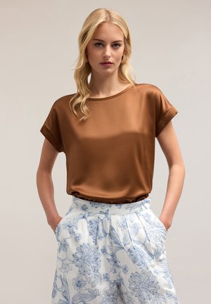 Braune Satinbluse mit kurzen Ärmeln und rundem Ausschnitt, kombiniert mit weißen, blumengemusterten Hosen, die seitliche Taschen und Falten haben.