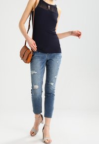 Marinblå ärmlös topp med genomskinliga detaljer vid halsringningen, matchad med slitna blå jeans och grå klacksandaler. Brun axelväska.