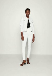 Bela denim jakna z srebrnimi gumbi, črna oprijeta majica in bele skinny jeans. Model nosi črne sandale na visoki peti. Preprost dizajn.