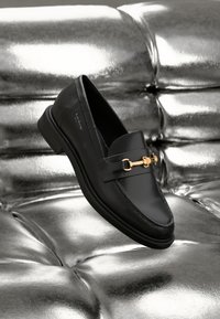 Schwarze Leder-Loafer mit goldfarbener Hardware. Der Schuh zeichnet sich durch ein elegantes Design und eine glatte, glänzende Oberfläche aus.