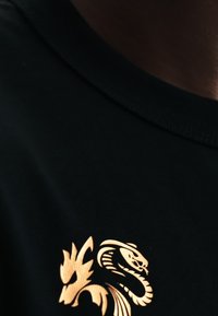 Camiseta negra con un emblema dorado de dragón y serpiente impreso cerca del escote en el pecho.