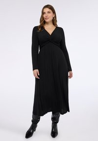 Černé šaty s dlouhým rukávem s uzlem na prsou, přiléhavým živůtkem a splývavou sukní; nošené s lesklými černými kozačkami nad kolena.