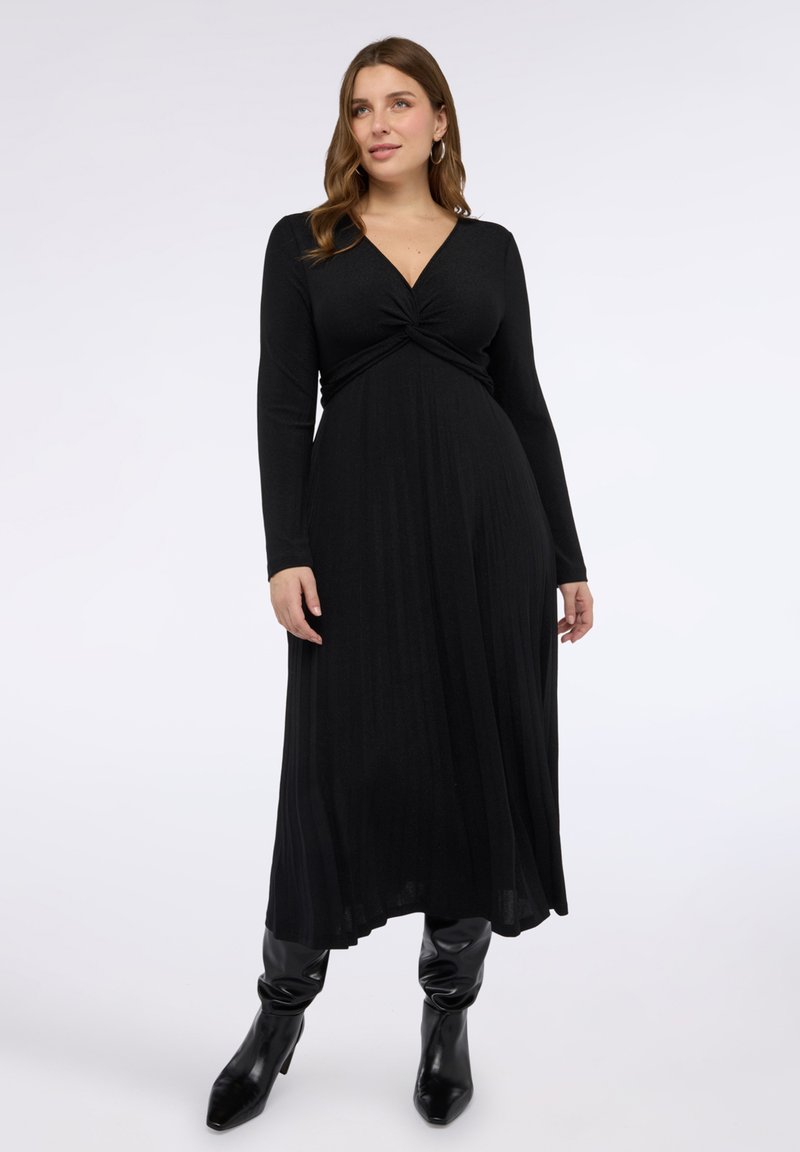 Černé šaty s dlouhým rukávem s uzlem na prsou, přiléhavým živůtkem a splývavou sukní; nošené s lesklými černými kozačkami nad kolena.