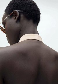 Sonnenbrille mit braunen Rahmen auf dem Gesicht eines Modells, das eine glatte Hauttextur und einen hellfarbenen, eng anliegenden Kragen am Nacken zeigt.