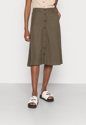 Femme portant une jupe midi vert olive boutonnée devant, un haut beige et des sandales blanches ouvertes à l'avant avec semelles noires, debout sur un sol gris.