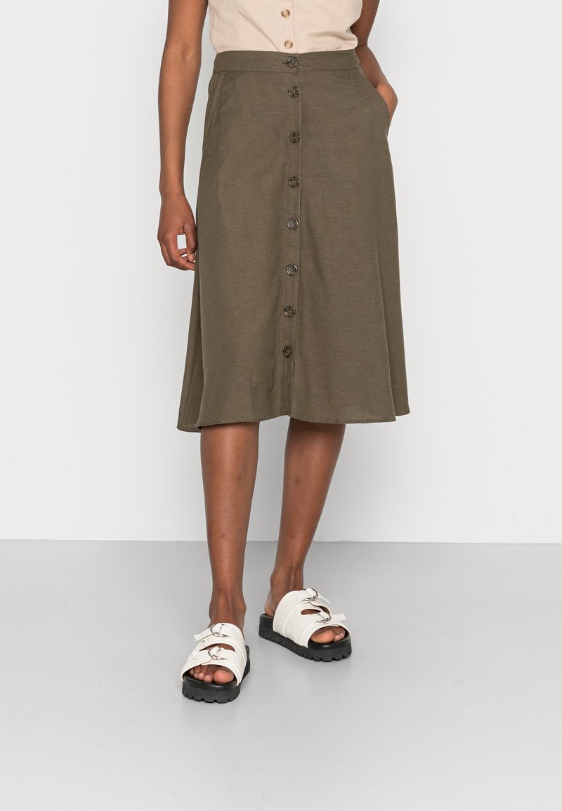 Femme portant une jupe midi vert olive boutonnée devant, un haut beige et des sandales blanches ouvertes à l'avant avec semelles noires, debout sur un sol gris.