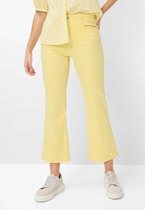 Parte inferiore del corpo di una persona che indossa pantaloni gialli a vita alta, a zampa e cropped, una maglietta gialla a maniche corte con trama e scarpe da ginnastica beige.