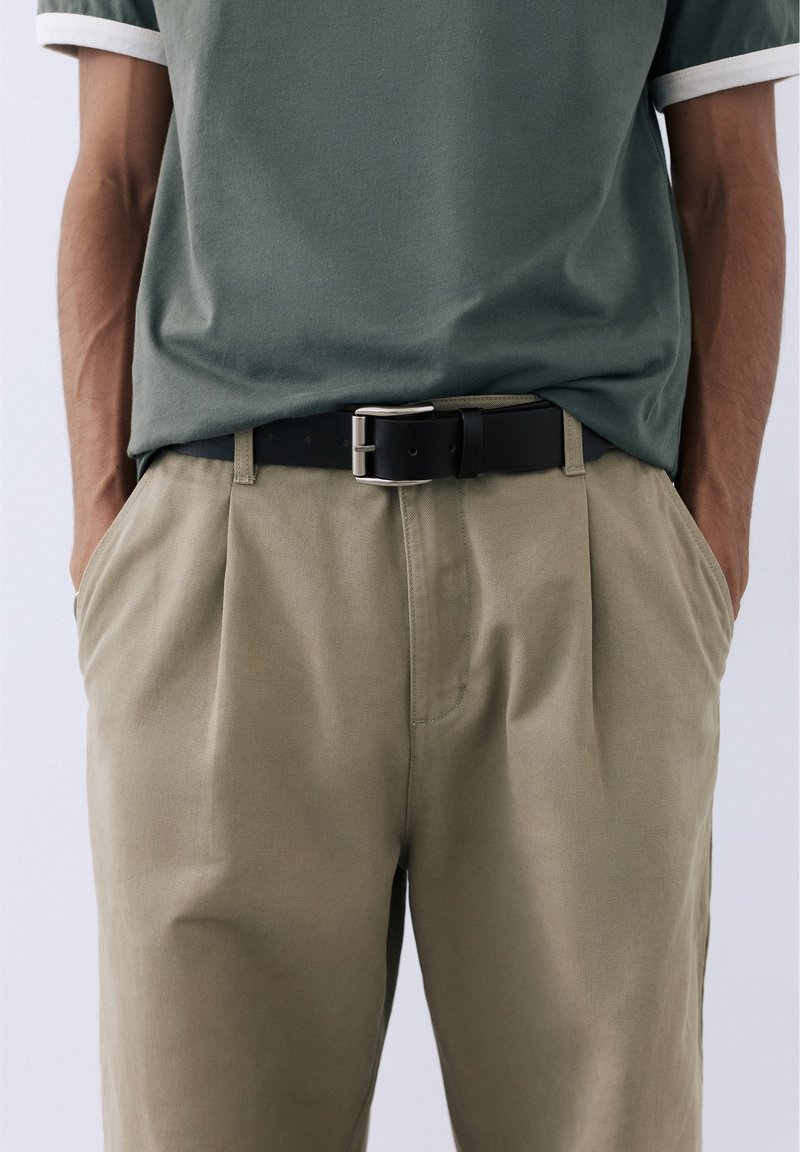 Personne portant un t-shirt vert sauge rentré dans un pantalon beige avec une ceinture noire, les mains reposant dans les poches du pantalon, sur un fond clair uni.