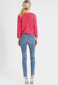 Blusa roja de flores con mangas largas, combinada con jeans ajustados azul claro y tacones altos plateados. La vista trasera muestra la textura de la blusa y el ajuste de los jeans.