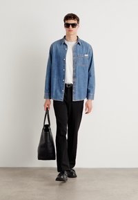 Blauwe denim overhemd met knopen, witte T-shirt, zwarte broek, zwarte schoenen en grote zwarte totebag. Model draagt donkere zonnebril.