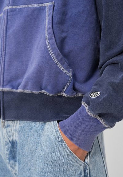 Hooded majica u gradijentnim nijansama plave boje. Ima rebraste manžete, bočne džepove i kontrastne šavove. Mekani teksturirani materijal.