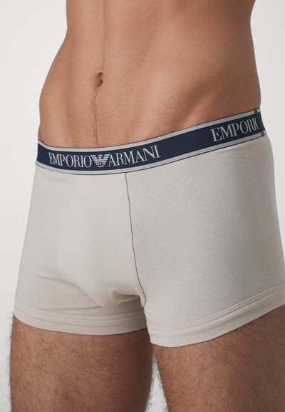 Beige bomullsboxer med en marinblå elastisk midja som har "Emporio Armani"-logotypen i vitt. Slät textur, medelhög design.