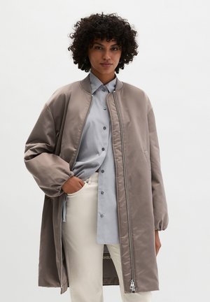 Vrouw met krullend haar, gekleed in een taupekleurige, lange bomberjas over een lichtblauw overhemd en crèmekleurige broek, staand met één hand in haar zak.