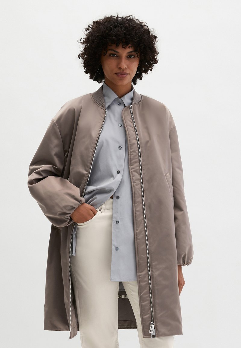 Woman with curly hair wearing a taupe long bomber jacket over a light blue button-up shirt and cream pants, standing with one hand in pocket.