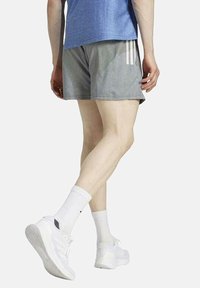Pantalones cortos deportivos grises con tres franjas blancas, de corte holgado, combinados con una camiseta azul y zapatillas blancas, mostrando una tela suave y un corte hasta la rodilla.