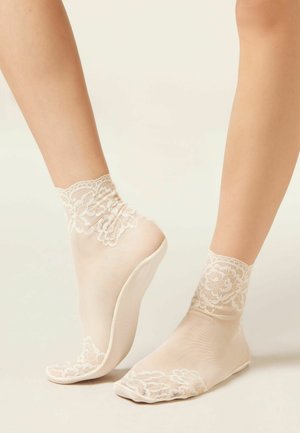 Piedi con calzini trasparenti beige alla caviglia con motivi floreali in pizzo su polsini, punte e talloni su uno sfondo chiaro e uniforme.