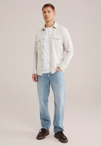 Joven de pie con la mano en el bolsillo, vistiendo una chaqueta blanca, una camiseta blanca, jeans de un azul claro y zapatos marrón oscuro sobre un fondo claro.