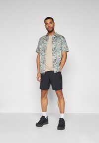 Kortermet mønstrete skjorte i blå, beige t-skjorte, mørke shorts og svarte joggesko med hvite sokker. Har tropisk-inspirerte grafikker.