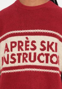 Maglione rosso e crema con testo "ISTRUTTORE APRÈS SKI", colletto rotondo, lavorazione a maglia testurizzata e motivi a bande orizzontali.
