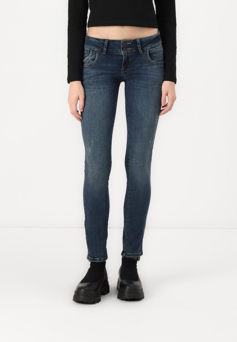 Femme portant un jean skinny bleu foncé avec des détails délavés, un crop top noir à manches longues et des bottines noires épaisses à plateforme sur fond blanc.