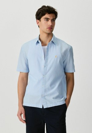 Férfi világoskék, rövid ujjú, piros csíkos gombos ingben és sötét nadrágban, egy kezét a zsebében tartva, egyszínű háttér előtt áll.