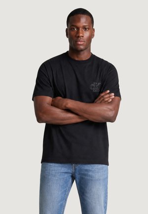 Mężczyzna o ciemnej karnacji, ubrany w czarny t-shirt z krótkim rękawem i małym logo "cK" oraz jasnoniebieskie jeansy, z założonymi rękami, neutralne tło.